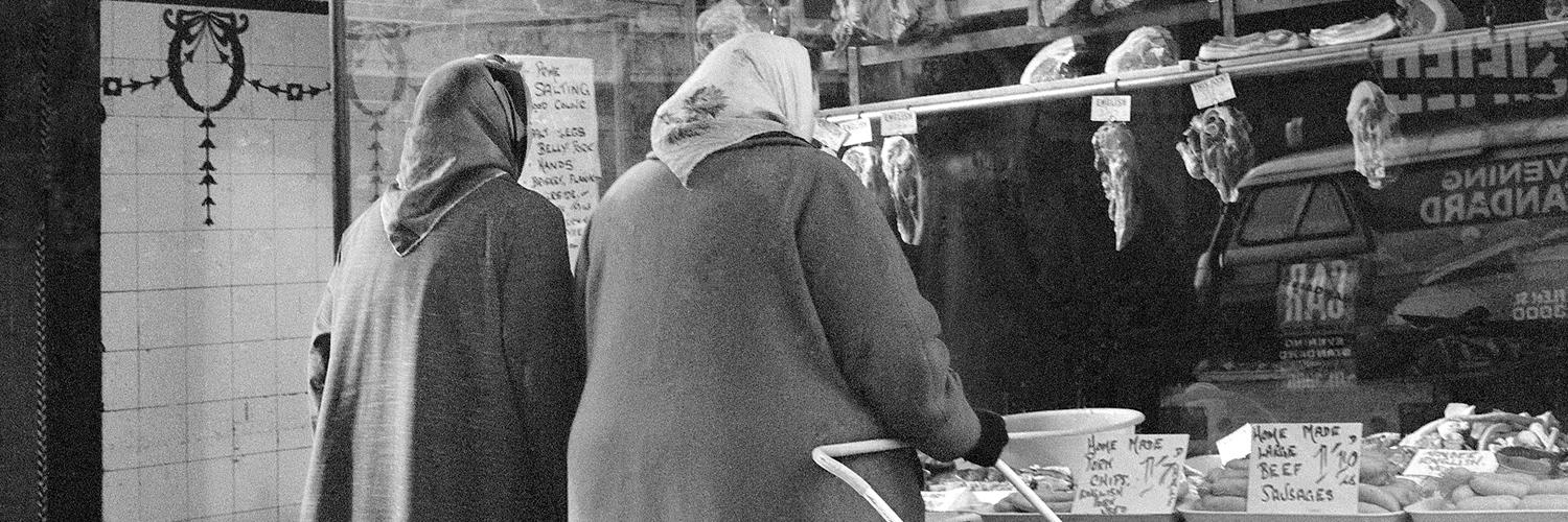 Black and white photo of two older women with headscarves on, one with a shopping trolly, standing outside what was then a butchers' window and is now the Young Vic box office entrance.