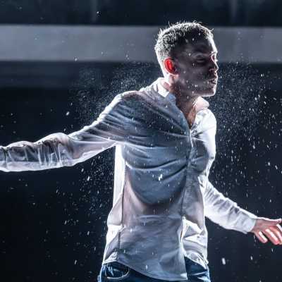 A man lit from behind, wearing a white shirt and blue jeans is standing on a stage. His arms are spread out to the side, and his eyes are closed. He is mid-action and there are sprays of water emanating from him. 