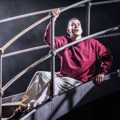 A man wearing a burgundy sweatshirt, grey tracksuit bottoms and black trainers sits on a metal bridge. His right hand is holding onto the railing above his hand, and his left hand is holding on to the bottom of the railing. He is looking up with eyes wide, and a slightly parted mouth.  