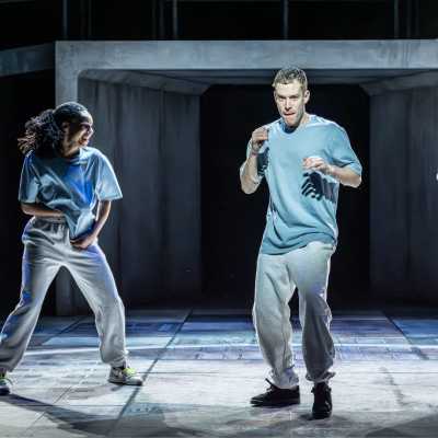 Two people wearing blue t-shirts, grey tracksuit bottoms and trainers stand on a stage, mid-action as if they were preparing for a boxing match. The woman on the left is laughing and the man in the middle is serious. 