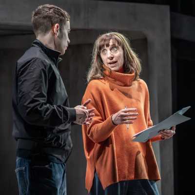 A man in a black jacket and jeans and a woman in an orange jumper and jeans are having a serious conversation on a stage. The woman is mid-speech and is holding a pen in her right hand and some sheets of paper in her left.  