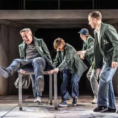 Four people stand on a stage wearing green school blazers. A fifth is sat on a school desk, with his legs upwards. Everyone is laughing. 