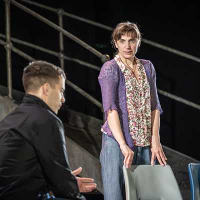 A man in black jacket is sat on a plastic chair. To his right, a woman in a purple cardigan and floral top stands, resting her hands on the back of a chair. She is staring intensely towards the camera. 