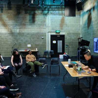 A group of people sat on chairs in a workshop. A person behind a desk presents something on a screen.