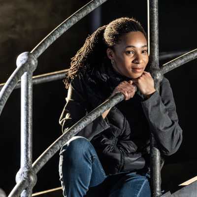 A young woman in a black jacket and blue jeans is sitting on a metal bridge. Her hands are gripping the railing in front of her, and her chin is resting on them. She appears to be deep in thought. 