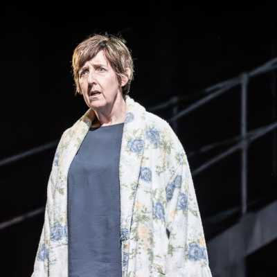 A woman in a floral dressing gown stands to the left of the image. She appears to be shocked and saddened.  