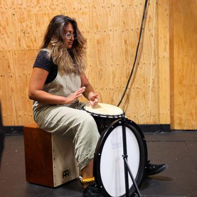 Musical Director Shakira Stellar is in the middle of clapping while sitting on a drum, holding an African drum between Stellar's legs and resting Stellar's foot on a drum pedal.