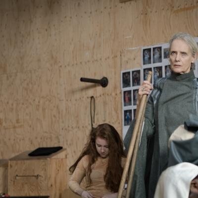 A female actor in long grey overalls stands solemn faced, holding a broom waiting for a scene to start © Leon Puplett