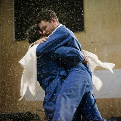Two male actors embrace one another whilst spinning on the spot as hay rains down all around them © Leon Puplett