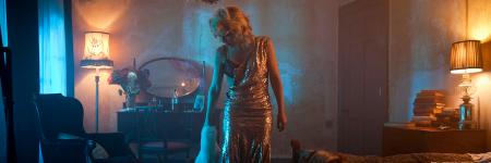 Gillian Anderson on set in the Young Vic short film, The Departure. She stands in a glittering dress, with one strap hanging off her shoulder in a hazy, darkly-lit room.