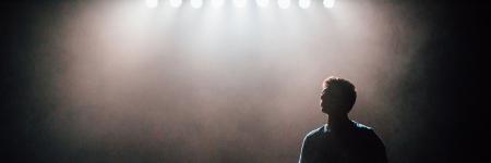 A silhouette of the head and shoulders of a young person looking out to the distance stands backlit onstage