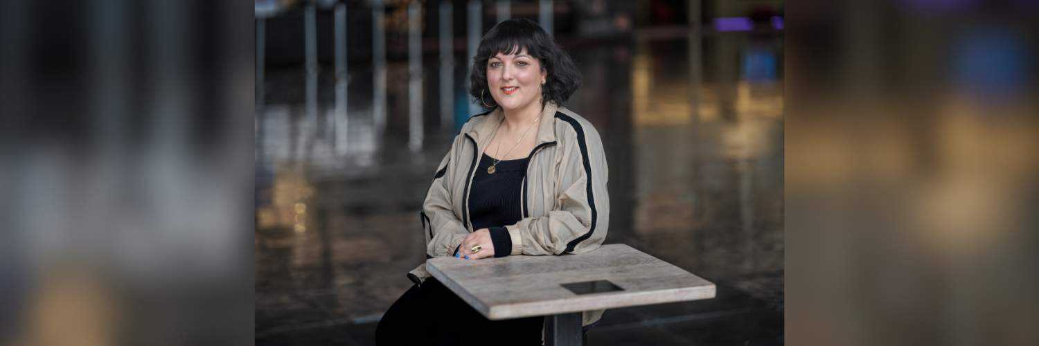 A light skinned, British South Asian woman with dark hair and a fringe, wearing a black top, beige jacket and gold jewellery, sat at a table