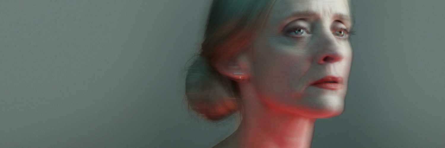 A blurred image of the head and shoulders of Anne-Marie Duff. Her blonde hair is pulled into a bun and her neck is lit from below with a red light. 