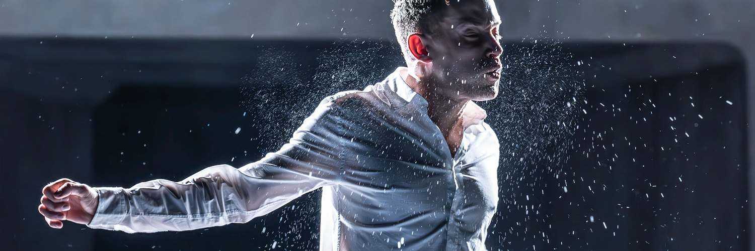 A man lit from behind, wearing a white shirt and blue jeans is standing on a stage. His arms are spread out to the side, and his eyes are closed. He is mid-action and there are sprays of water emanating from him