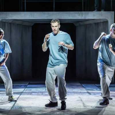 Three people wearing blue t-shirts, grey tracksuit bottoms and trainers stand on a stage, mid-action as if they were preparing for a boxing match. The woman on the left is laughing, the man in the middle is serious and the man on the right is worried. 