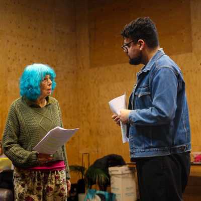 Two people standing with scripts in a rehearsal space with wooden walls. A seated person is visible blurred behind them.