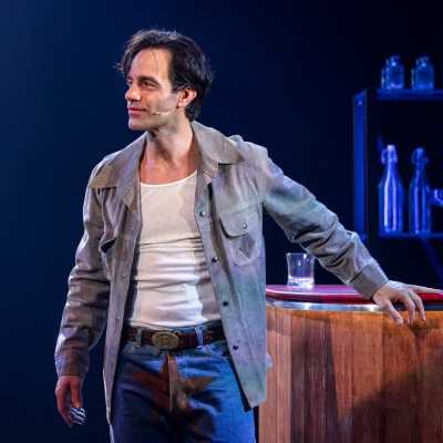 A clean-shaven person with dark hair stands in front of a bar, wearing a grey shirt and blue jeans with a suede star patch over the crotch.