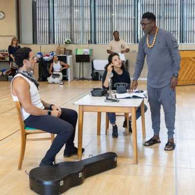 Director Kwame Kwei-Armah stands at a table, speaking to the show's leads  Anoushka Lucas and Ramin Karimloo, who are sitting down. They look at him, as he points to the script on the table. Various members of the cast and crew can be seen sitting and standing in the background.