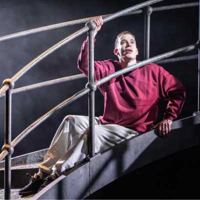 A man wearing a burgundy sweatshirt, grey tracksuit bottoms and black trainers sits on a metal bridge. His right hand is holding onto the railing above his hand, and his left hand is holding on to the bottom of the railing. He is looking up with eyes wide, and a slightly parted mouth.  