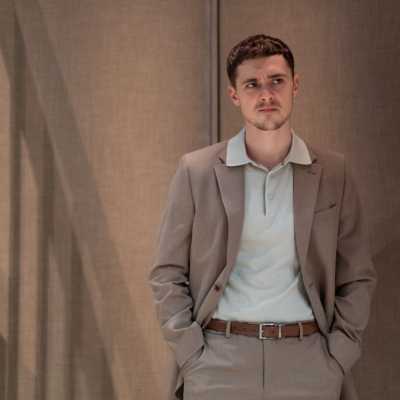 A white man with brown hair stands against a wall with his hands in his pockets. He is wearing a light brown suit and white shirt.