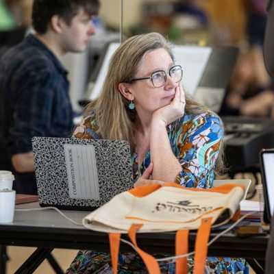 A solo shot of show playwright Sarah Ruhl. She is sat at a table, behind an open composition book, with her head resting in one hand, looking up to her left.