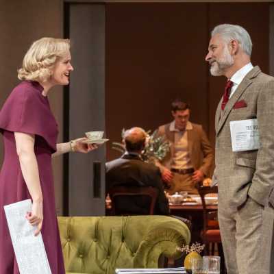 A tall female with blonde hair, wearing a purple dress and holding a teacup, faces towards a white man of average height, wearing a grey suit and holding a newspaper under his arm. They are having an animated conversation. Visible through a door behind them are some men sitting at a dining table. 