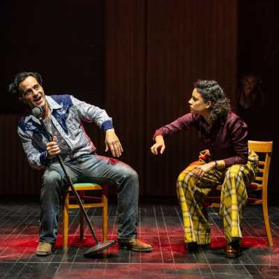 A light-skinned mixed race woman tries to grab an old-fashioned microphone out of the hands of a clean-shaven person with dark hair. Both are seated.