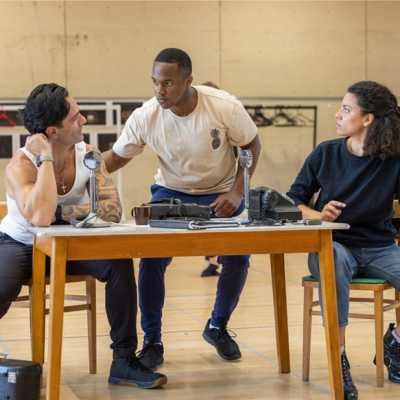 Cast members Ramin Karimloo, Durone Stokes, and Anoushka Lucas are gathered around a table. Ramin and Anoushka are looking at Durone, who appears to have just arrived to deliver some important news. 