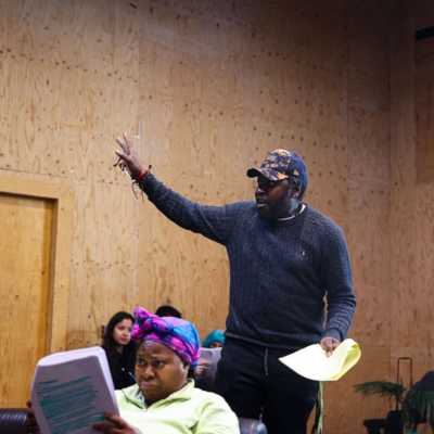 A person holding a script stands in a rehearsal room with wooden walls. One arm is raised in a peace sign. In front of him a person seated on a sofa with a script is visible. 