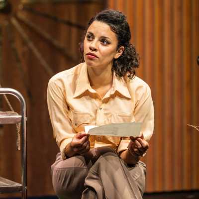 A light-skinned mixed race woman with freckles sits with legs crossed, holding a piece of paper. She wears a yellow shirt and light brown trousers.