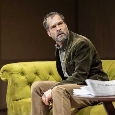 A white man of average height with short brown hair sits on a green sofa. He wears a green corduroy jacket, beige trousers and a bluey-green shirt. He looks slightly pale and unwell. 