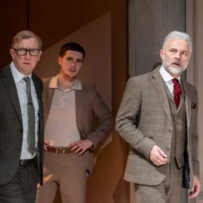 Three white men, all of average heights, wearing suits, peer around the corner of a door together. 