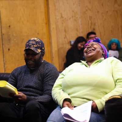 Two people sat on a sofa holding scripts in a rehearsal room with wooden walls. They are laughing. 