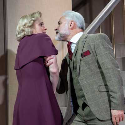 A white man with silver hair and a beard holds a tall female with blonde hair and green eyes by her arm, forcefully. They stand at the bottom of a staircase. He wears a grey suit and she wears a purple dress.