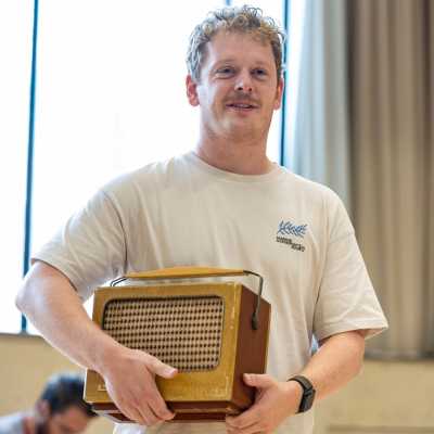 Cast member Chris Jenkins is pictured holding a 50s style radio. He is wearing a white t-shirt.