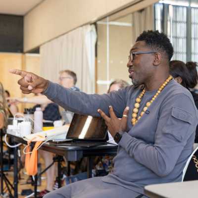 Director Kwame Kwei-Armah is pictured sitting down, pointing with his right hand at something or someone beyond the photograph. He is wearing a grey sweat-shirt with matching swaet pants, glasses and a necklance made of wood. We are given the impression he is directing cast members stood in front of him.