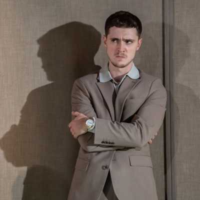 A white man with brown hair stands against a wall with his arms crossed over his chest. He is wearing a light brown suit, a white shirt and a smart wrist watch.  