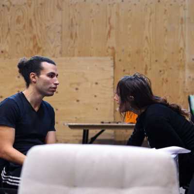 Two people sat facing each other in a rehearsal room with wooden walls. One person leans towards the other as if speaking intently. 