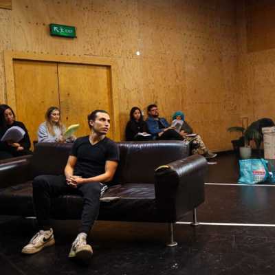 A person standing speaks to another person seated on a sofa in a rehearsal room with wooden walls. Seated people watching are visible in the background. 