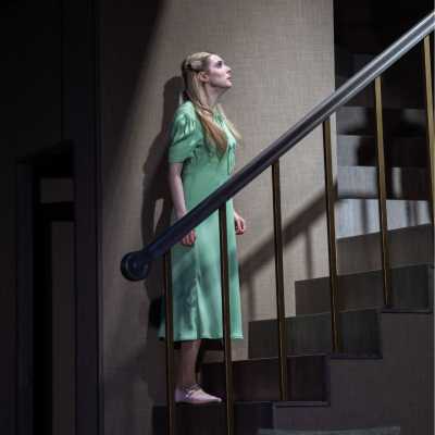 A petite, pale woman with chest-length blonde hair stands at the bottom of a staircase looking up. She is wearing a pale green dress. 