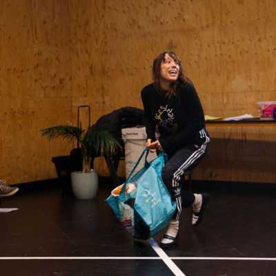 A smiling person crouches in a rehearsal room holding a large shopping bag.  