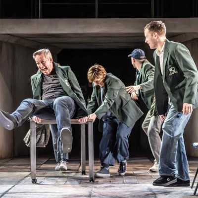 Four people stand on a stage wearing green school blazers. A fifth is sat on a school desk, with his legs upwards. Everyone is laughing.