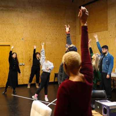 A Group of people standing in a circle in a studio with wooden walls raising one arm in the air