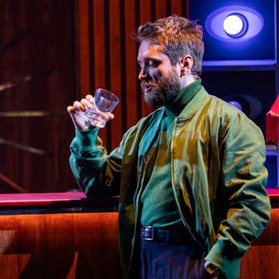 A white male with light brown hair and a beard wears a green jacket and is drinking from a glass