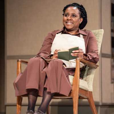 A Black woman with shoulder length dreadlocks pulled up in a bun wearing a brown dress and a white apron sits in a chair. She is smiling and holding a green book in her hands. 