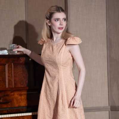 A petite, pale woman with chest-length blonde hair stands next to a piano. She is setting a glass of water on top of it but looking the other way. She wears a pale pink dress with frilly shoulders. 