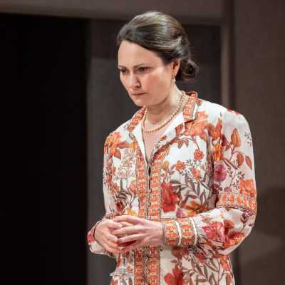 A tall woman with brown hair wearing a flower patterned pink and orange dress stands looking thoughtful. She is holding a wine glass in both hands.