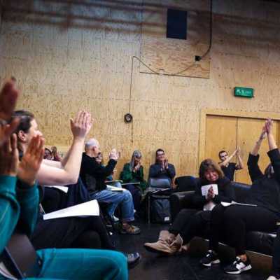 A group of people seated in a rehearsal room with wooden walls clapping. 