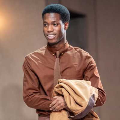 A Black man of average height with short Afro hair wearing a brown shirt and tie and holding a tan jacket stands smiling. 