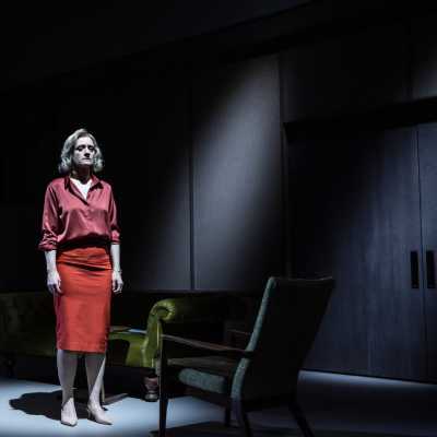 A female with blonde hair stands alone on the stage. She is wearing a red skirt and a red satin shirt. Her arms are by her sides and she stares into the distance. 